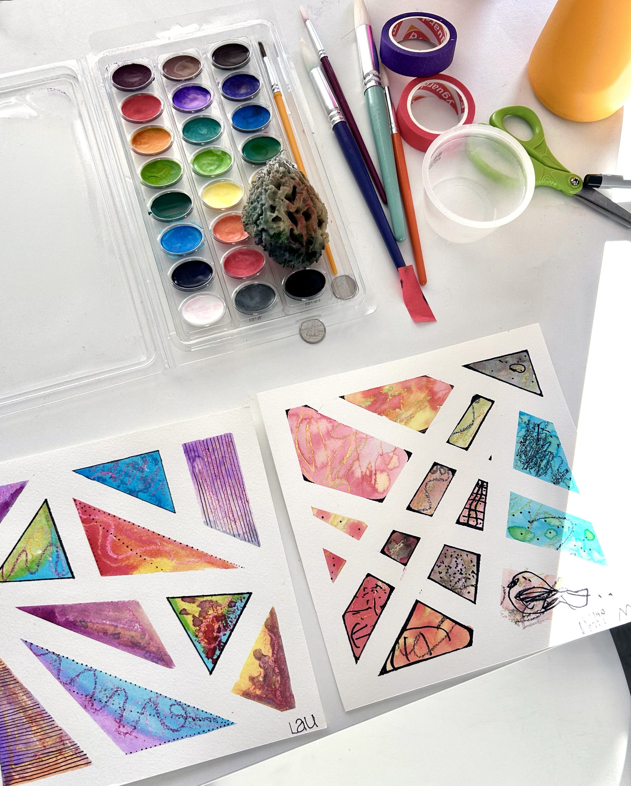 Compositions géométriques à l’aquarelle, palettes et pinceaux disposés sur une table.
