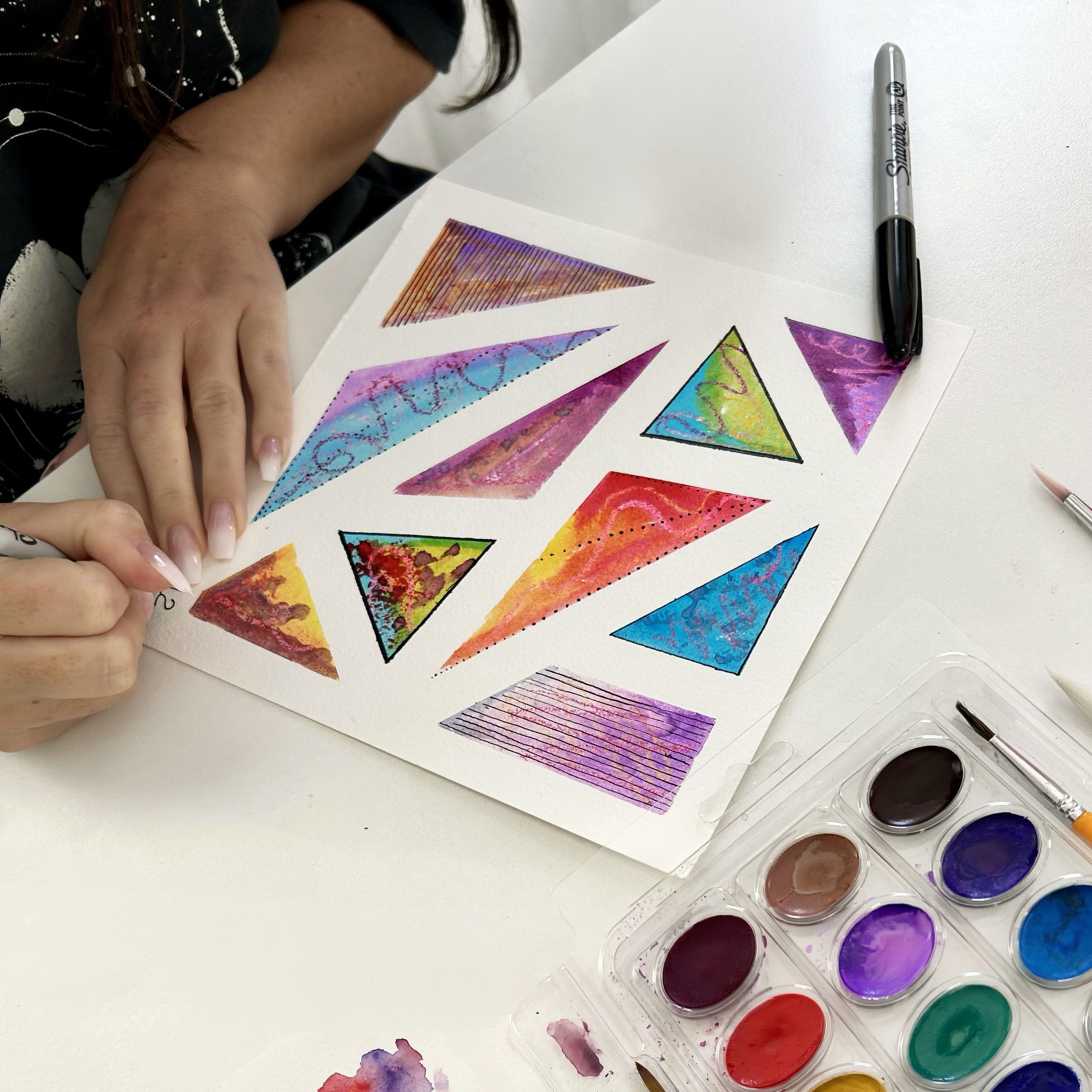 Des mains retirent le ruban adhésif d’une composition de triangles peints à l’aquarelle.